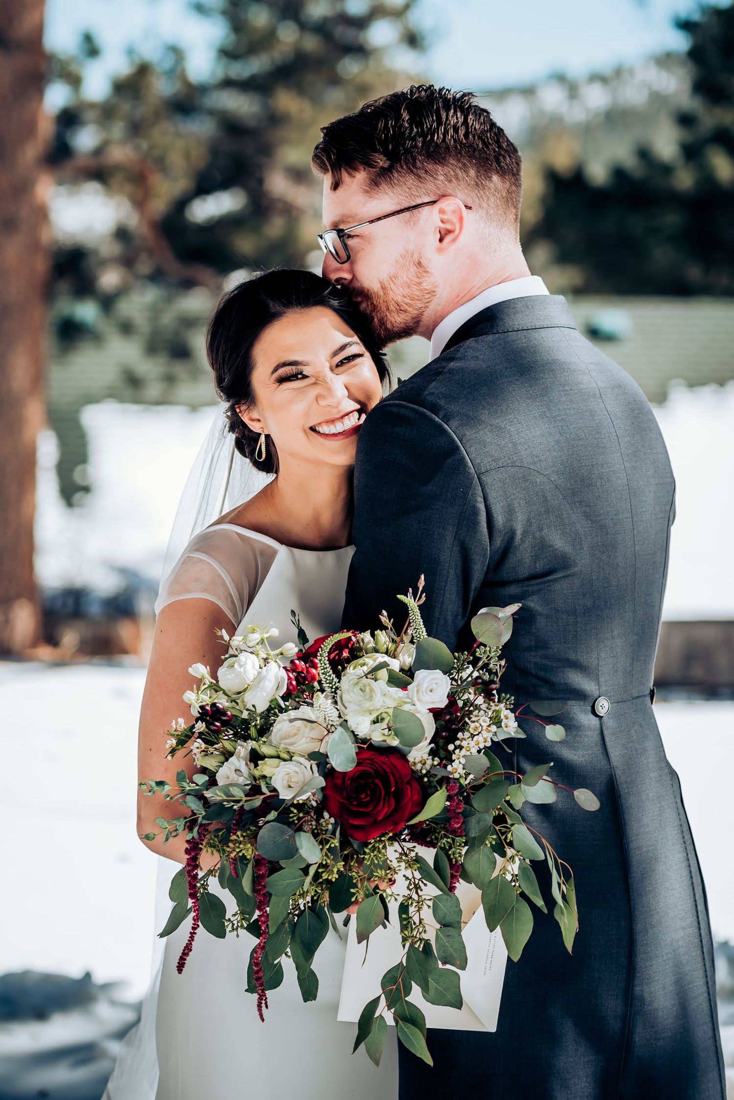 A joyful wedding couple embraces in a snowy mountain setting while holding a luxurious red and white bouquet.
