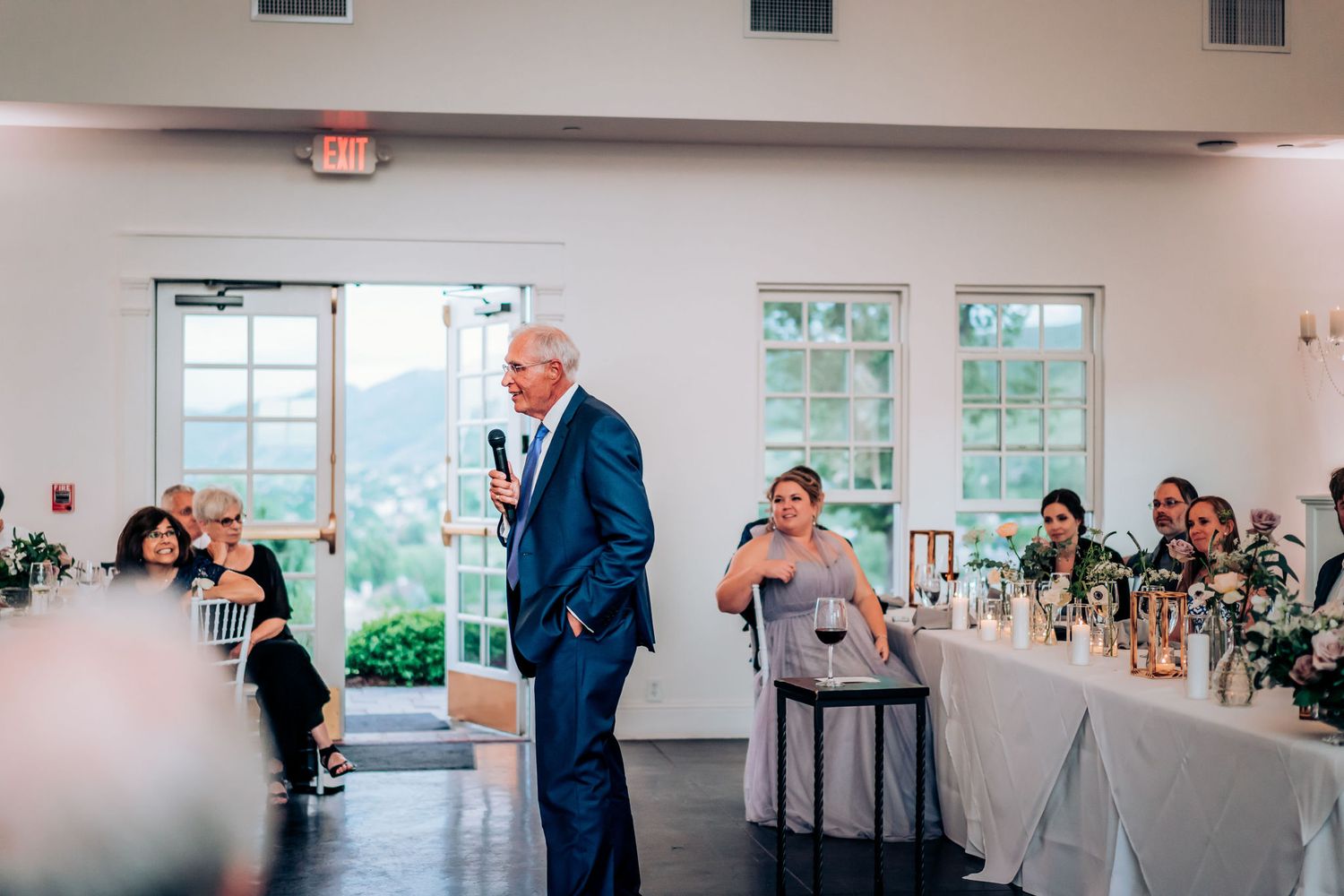 A wedding reception speech being delivered in an elegant white indoor venue with large windows.