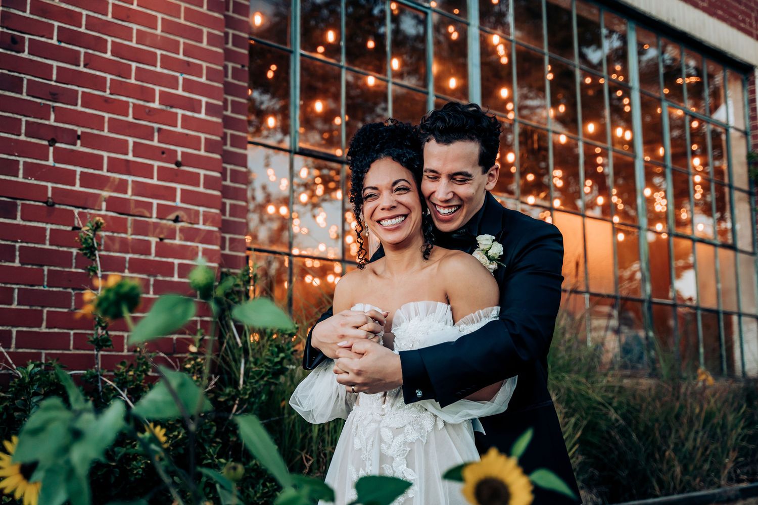Candid wedding photo showing laughter and joy against a brick wall with twinkling lights.