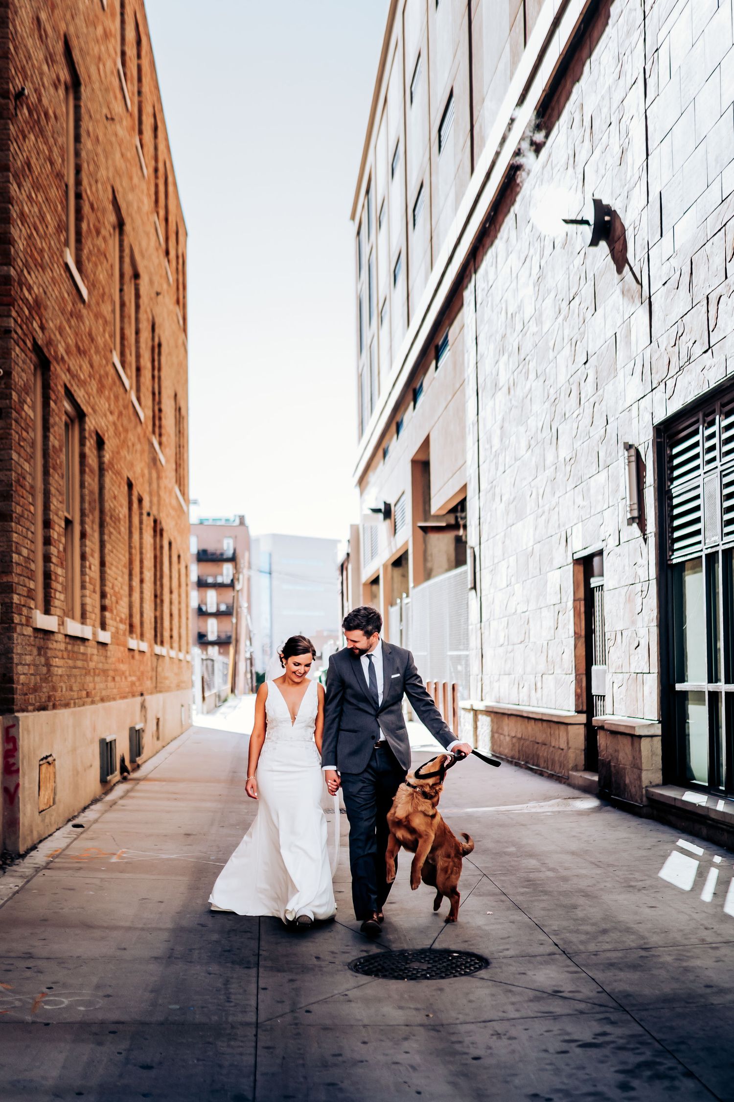 Wedding couple walks their excited dog down urban alley between brick and white buildings.