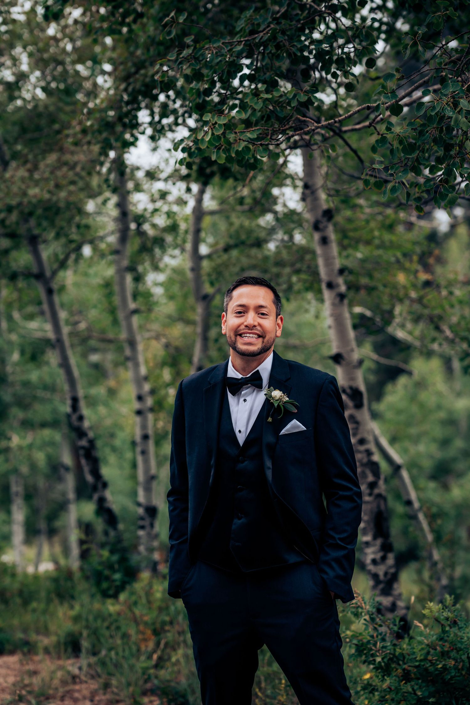 A groom in a navy tuxedo poses in front of an aspen forest on his wedding day.
