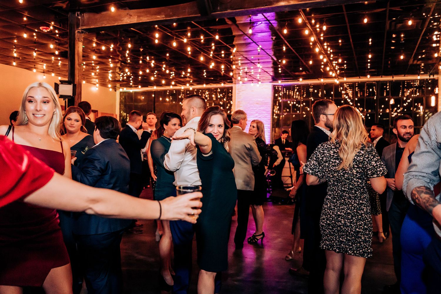Energetic crowd dancing under warm string lights at an indoor celebration.