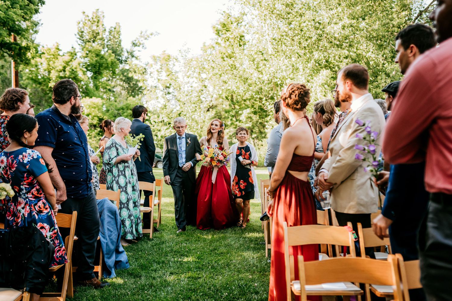 Outdoor wedding ceremony with guests seated on wooden chairs and a red dress standing out in the crowd.
