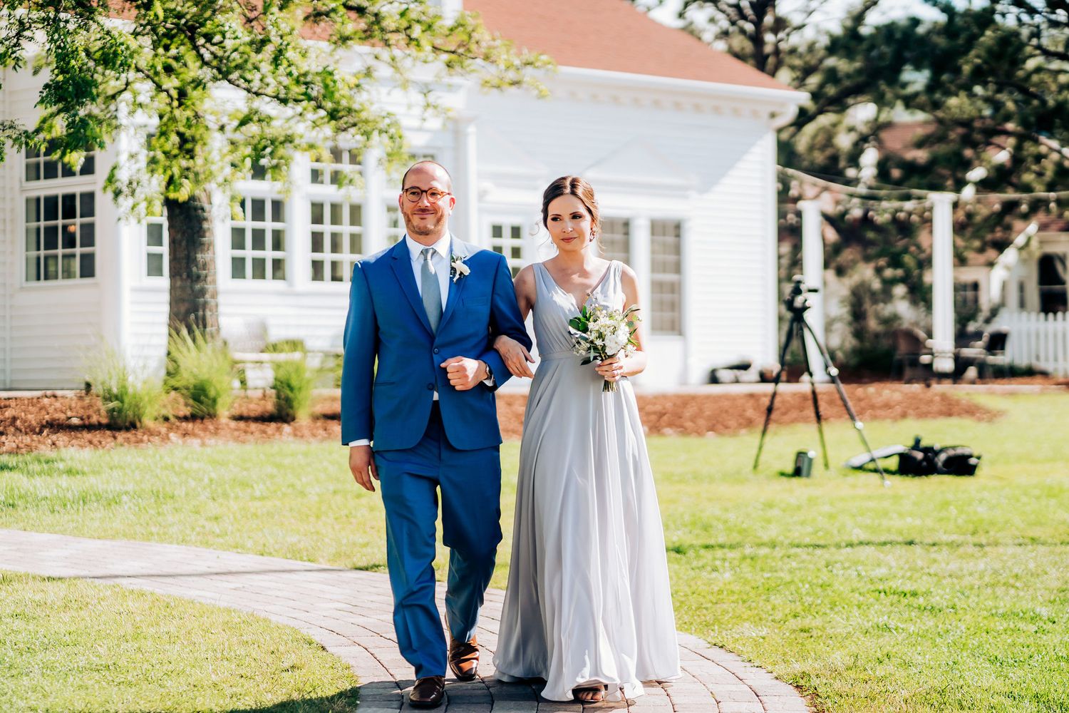 Wedding couple walks down garden path at elegant white venue on sunny day.