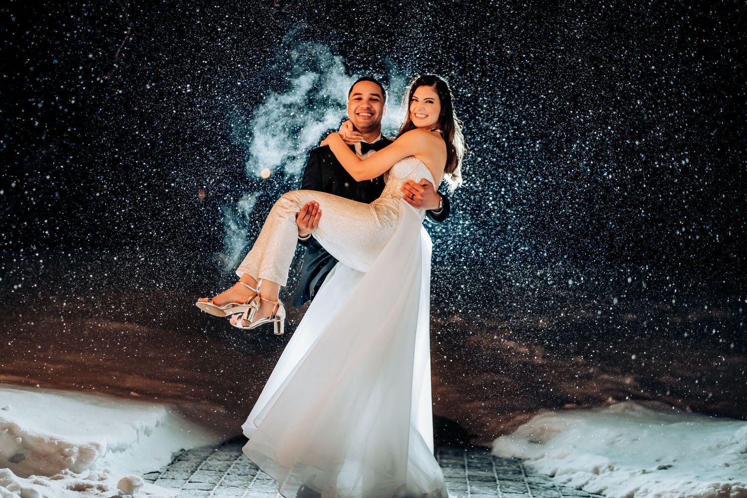 Wedding couple poses playfully in falling snow with ethereal lighting.