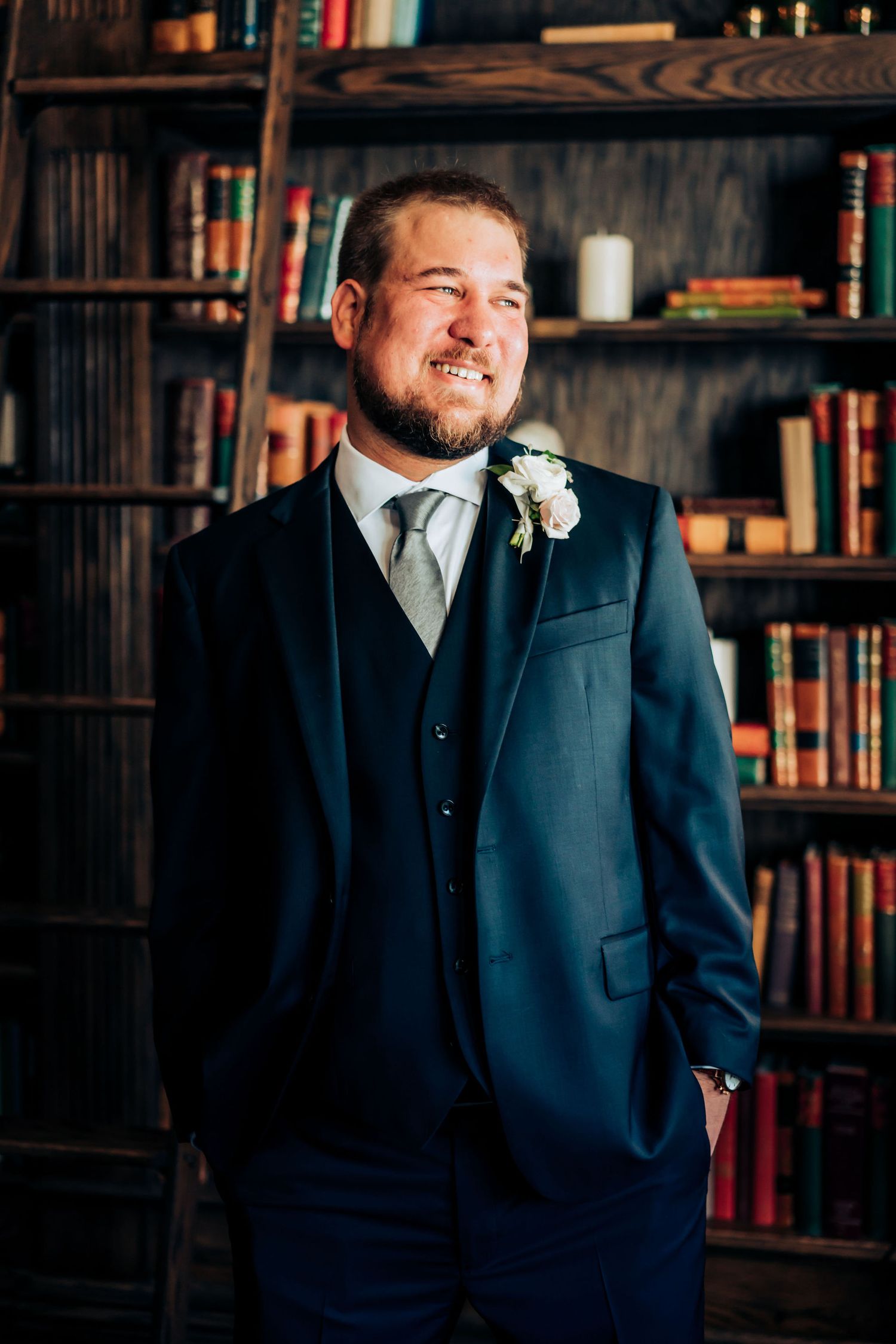 Person in navy suit and boutonniere poses in front of library bookshelf.