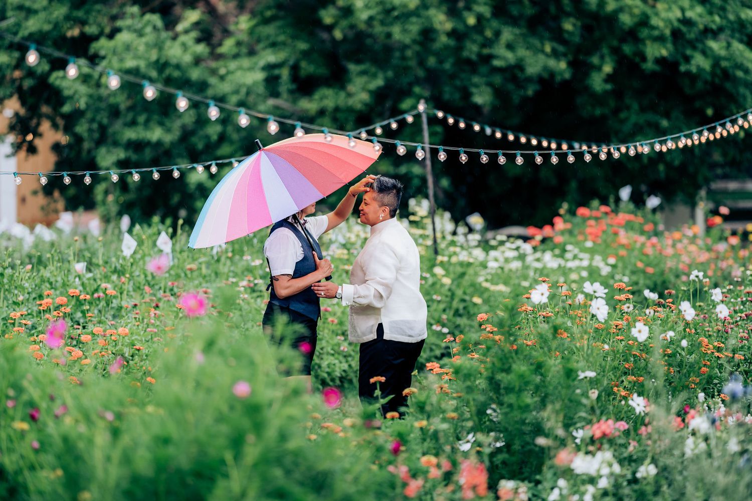 A romantic couple shares a colorful umbrella in a lush garden filled with flowers and string lights.