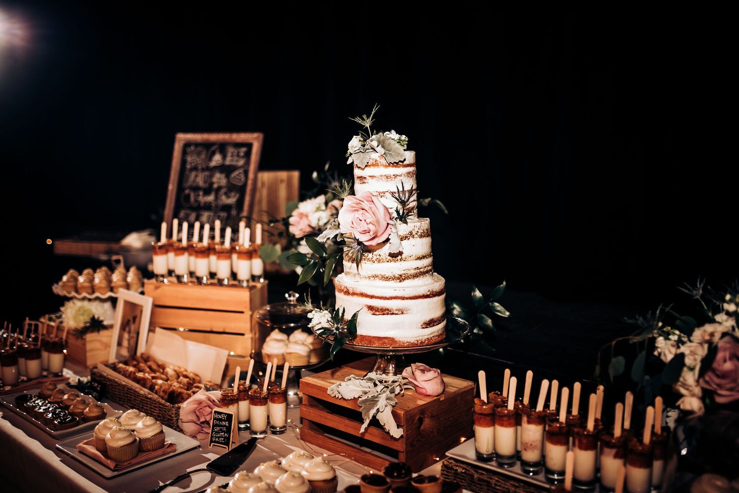 Rustic wedding dessert display with multi-tiered cake and illuminated treats on wooden stands.