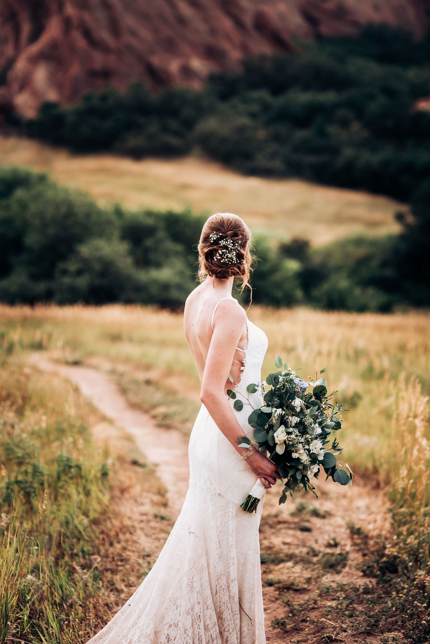 A bride in a fitted lace dress holds a greenery bouquet against red rock formations.