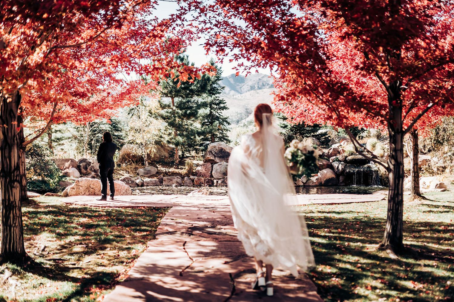 Wedding dress billowing in breeze under red maple trees in a scenic garden.