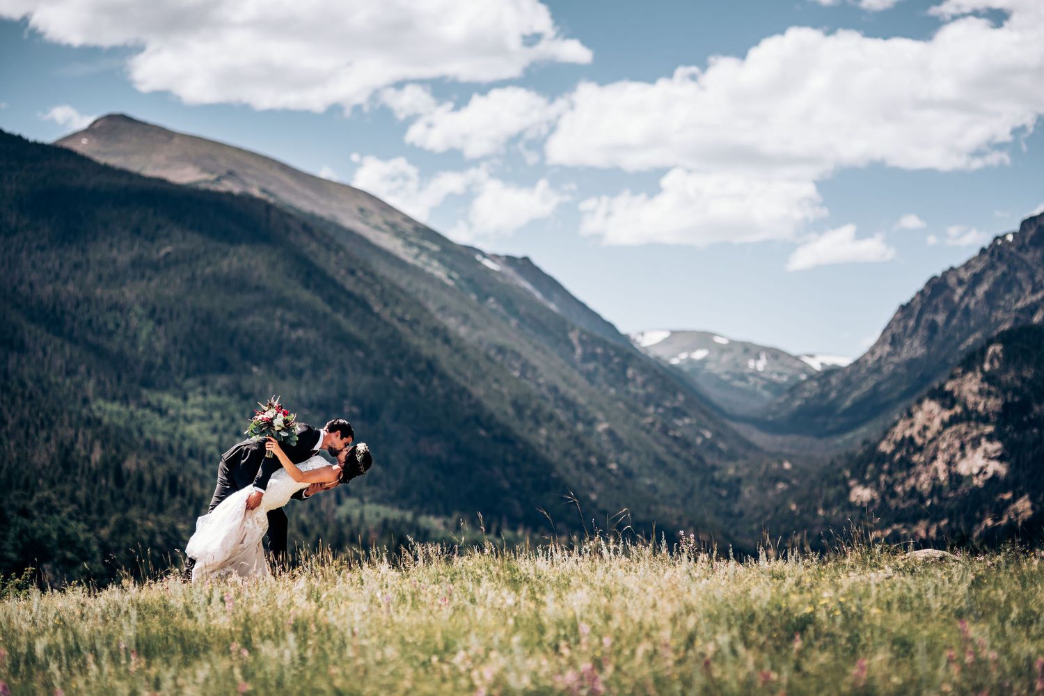 Wedding couple embraces in mountain meadow with dramatic valley and peaks in background.