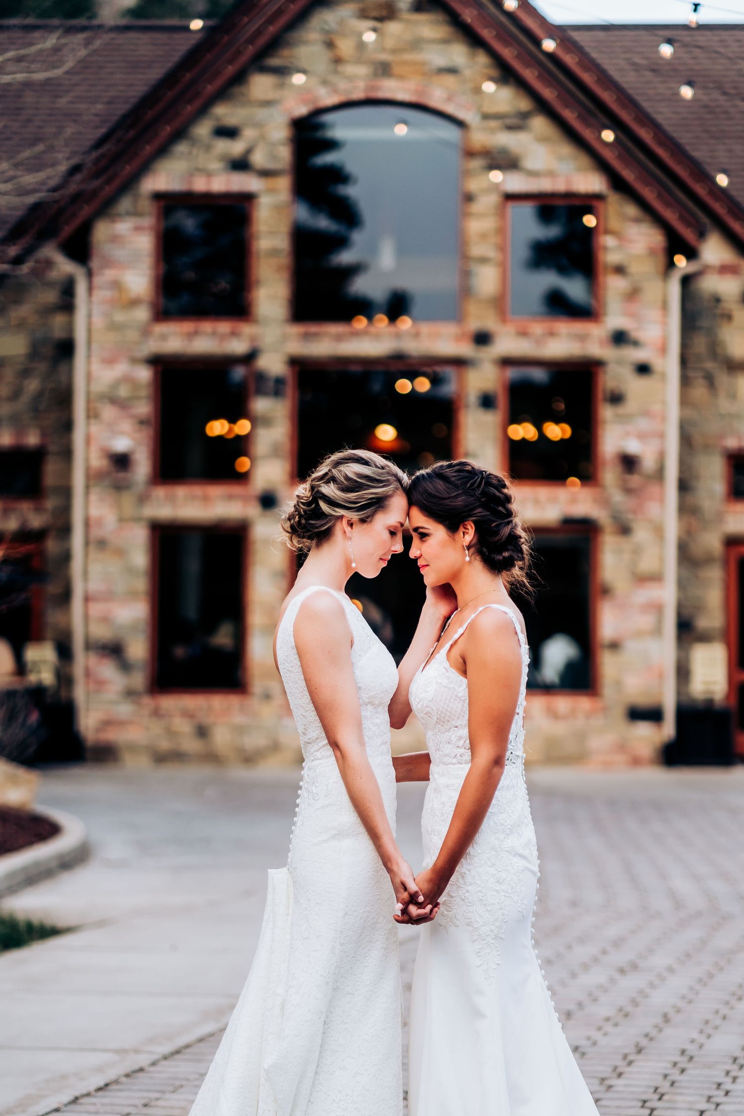 Two brides in elegant white wedding gowns share an intimate moment in front of a rustic stone venue with glowing lights.