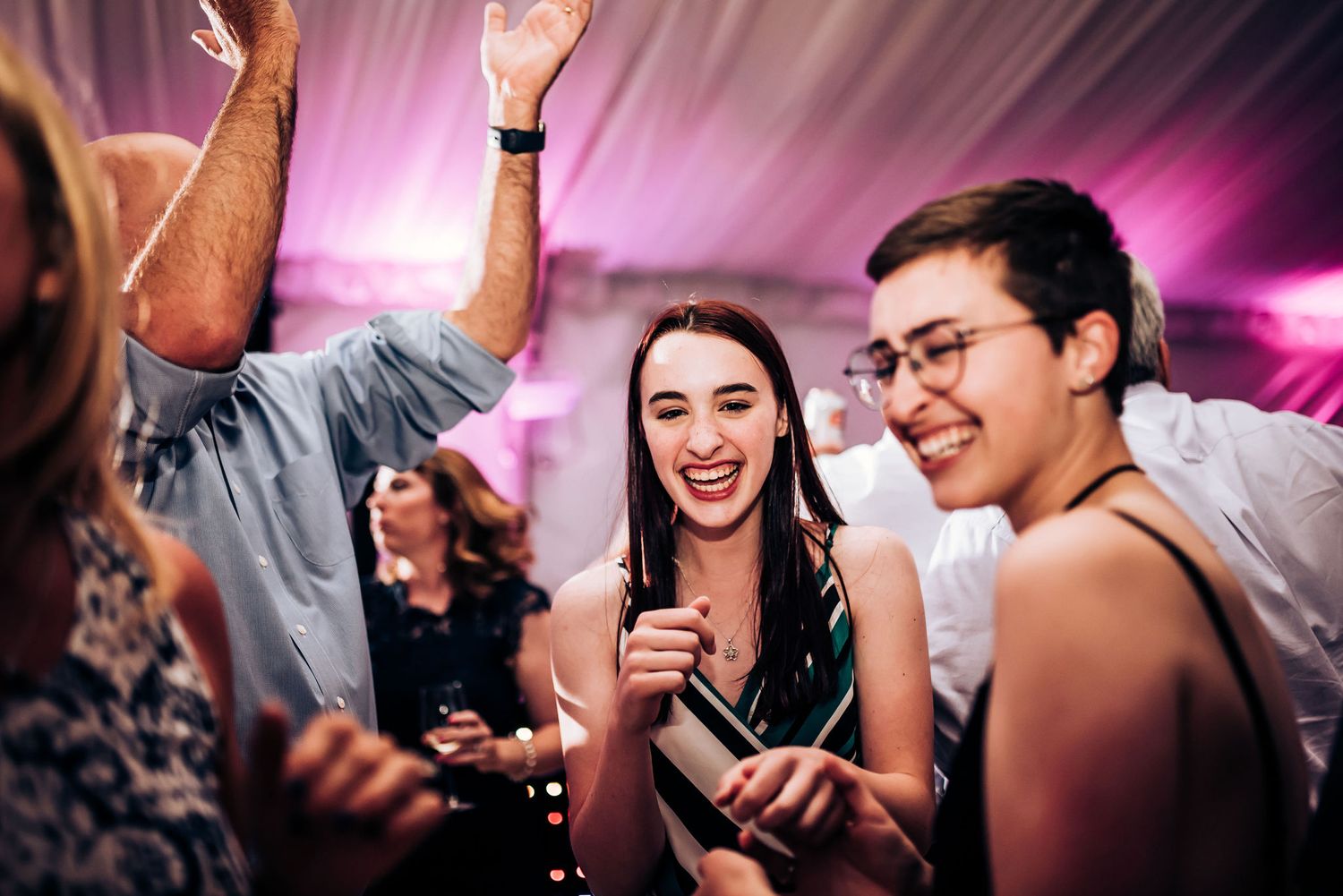 Energetic guests dance and laugh at a vibrant wedding reception with pink lighting.