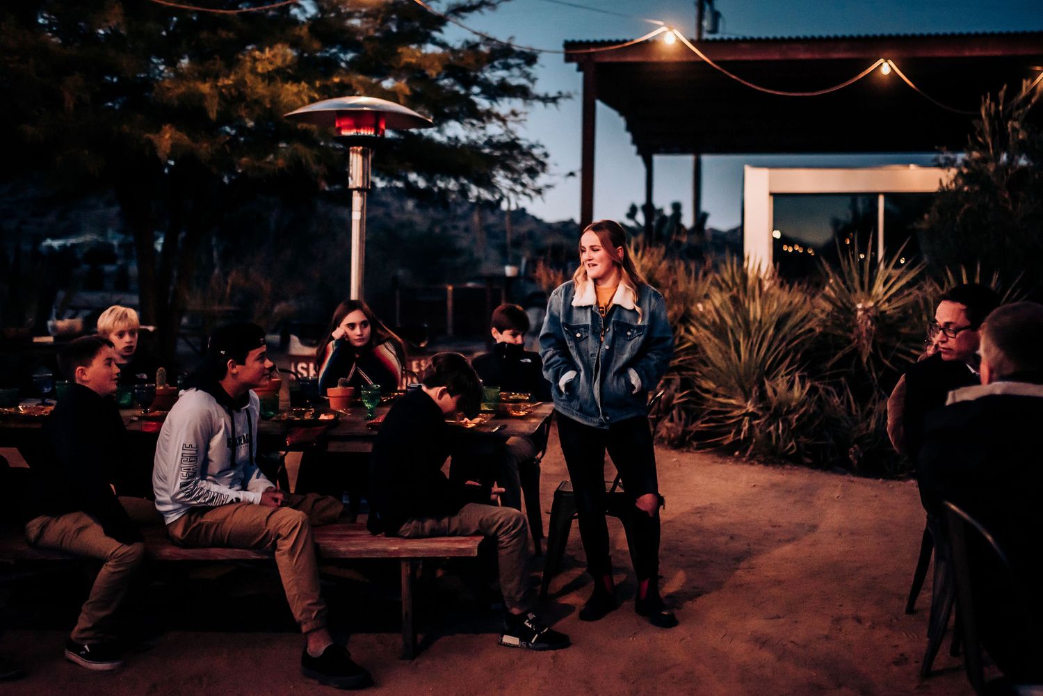Guests gather at an outdoor patio dining area with glowing heat lamps during twilight hours.