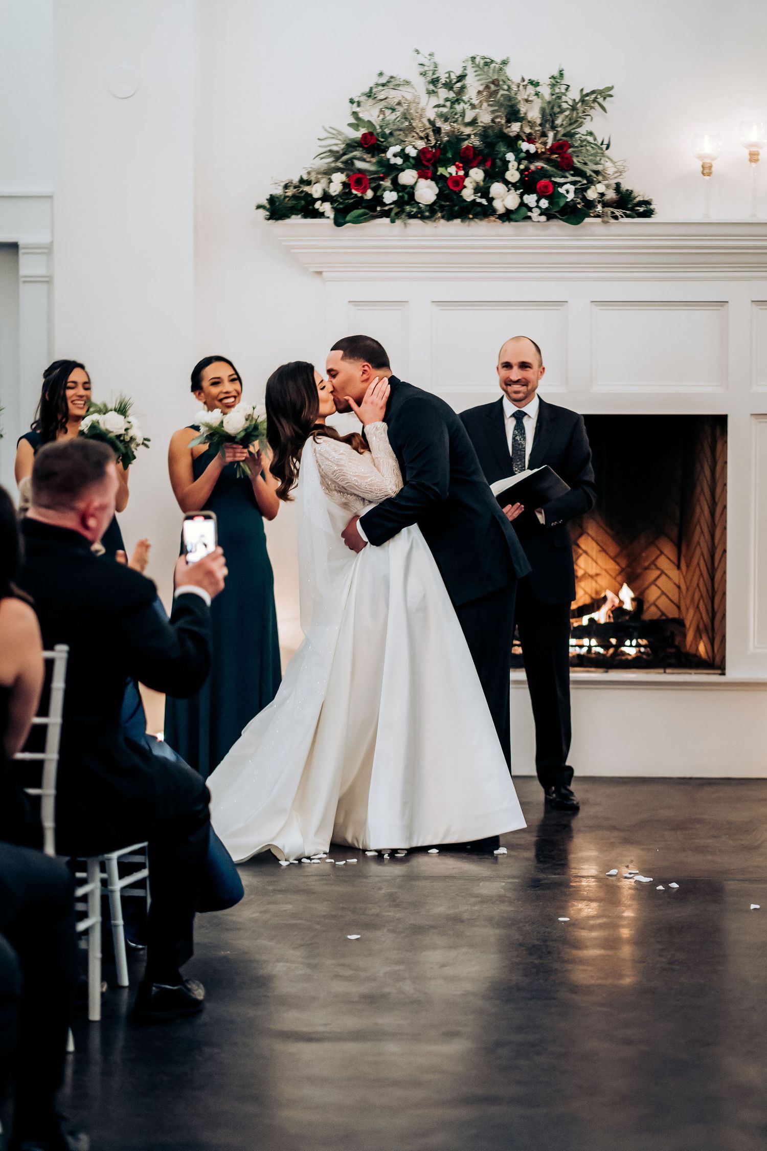 The newlyweds share their first kiss in front of a decorated fireplace with floral arrangements.