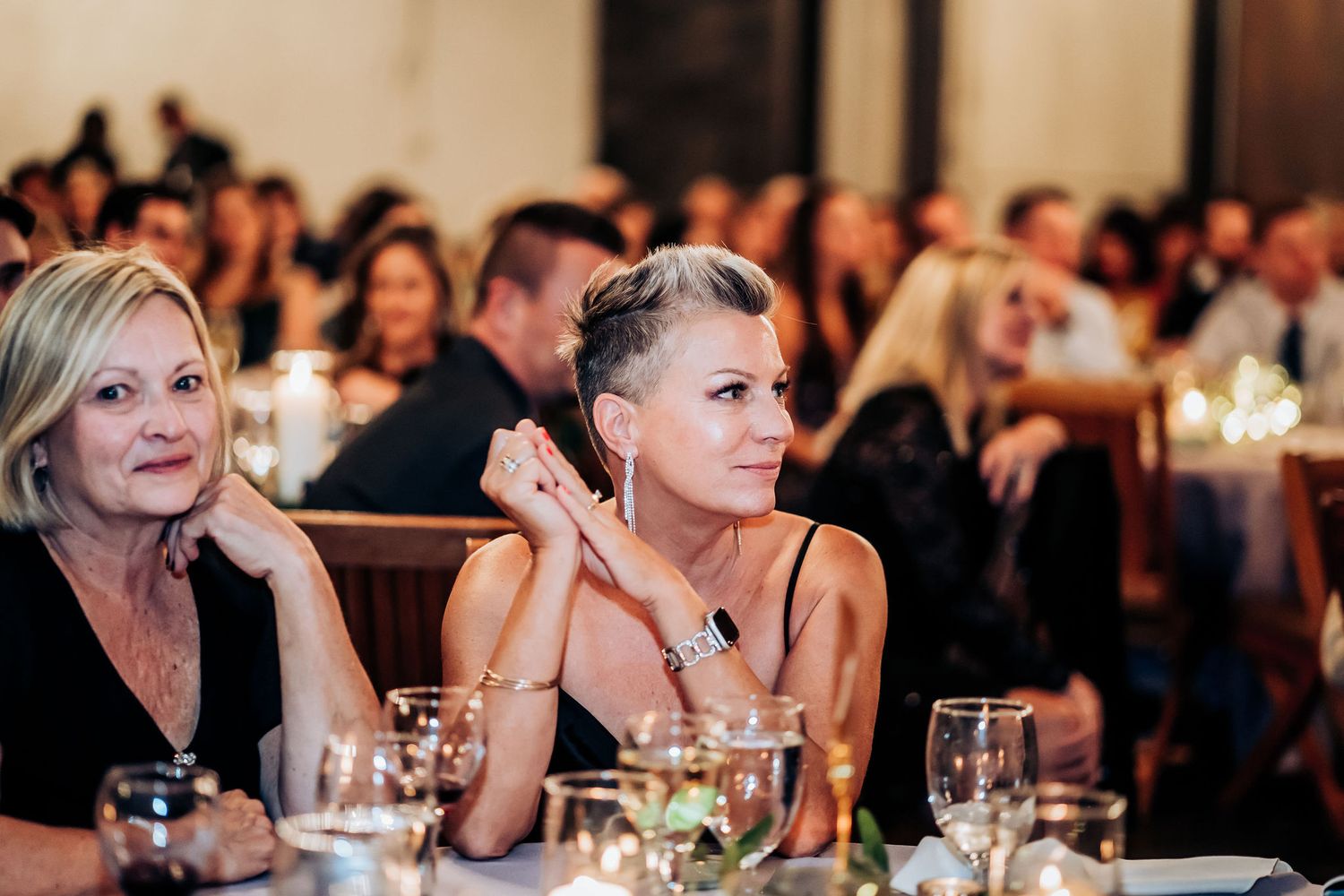 Wedding guests attentively watch speeches during an indoor reception at an elegant venue.