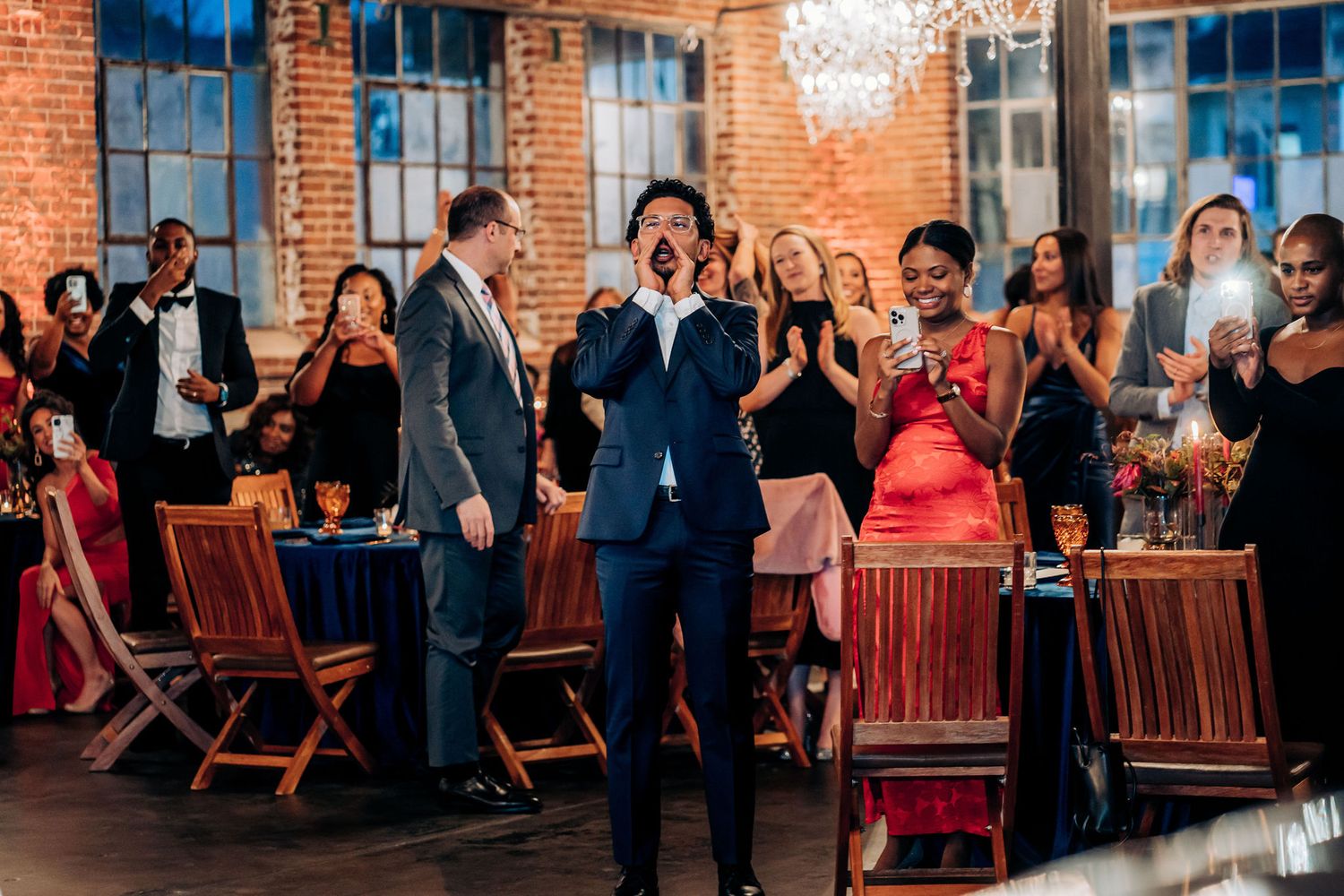 Wedding guests standing and celebrating during reception in rustic brick venue with chandelier.