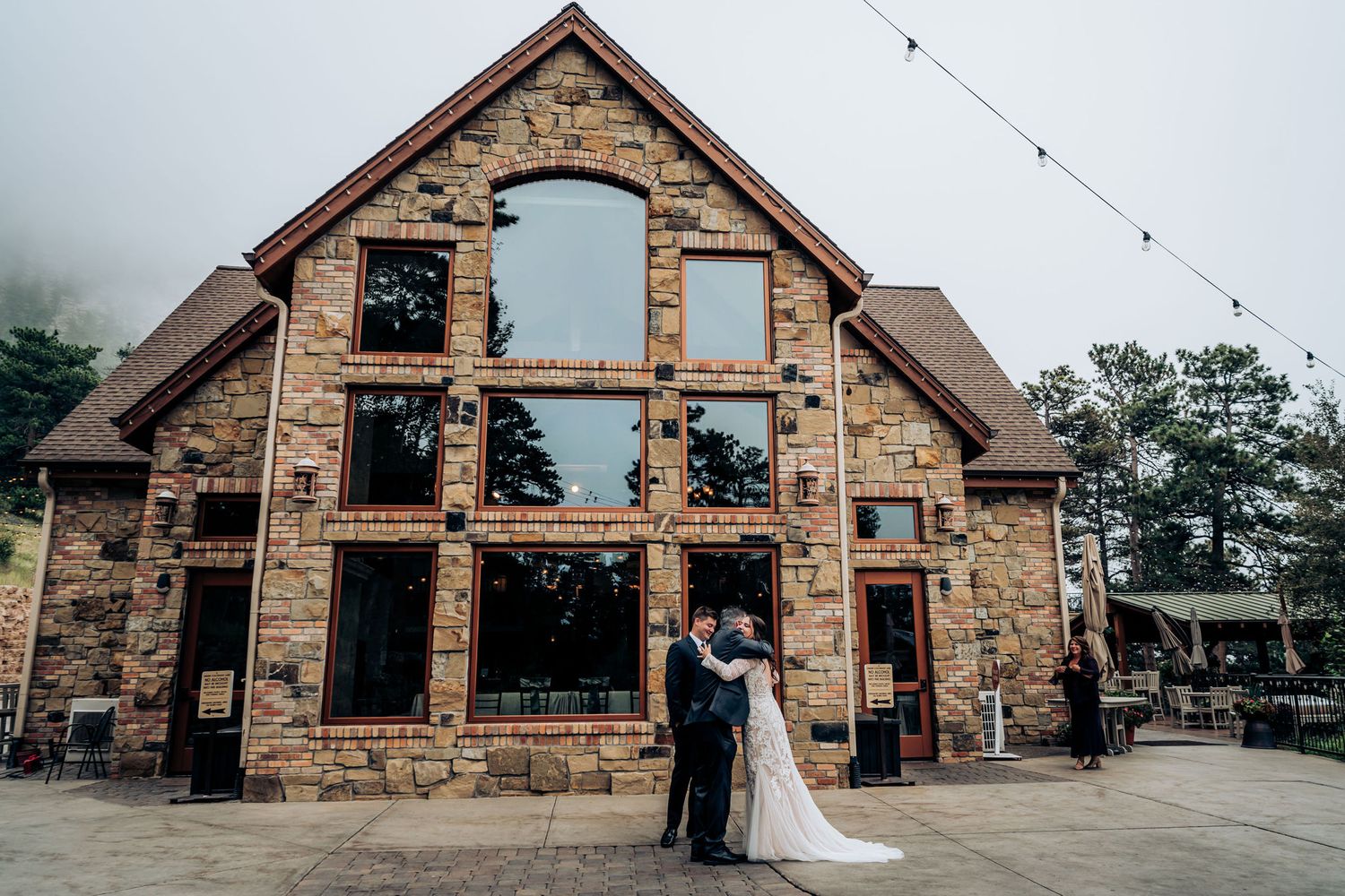 Wedding couple poses in front of stunning stone lodge with large windows and string lights.