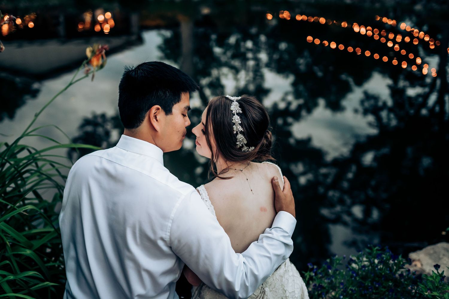 A couple embraces against twinkling lights and foliage during twilight hours.