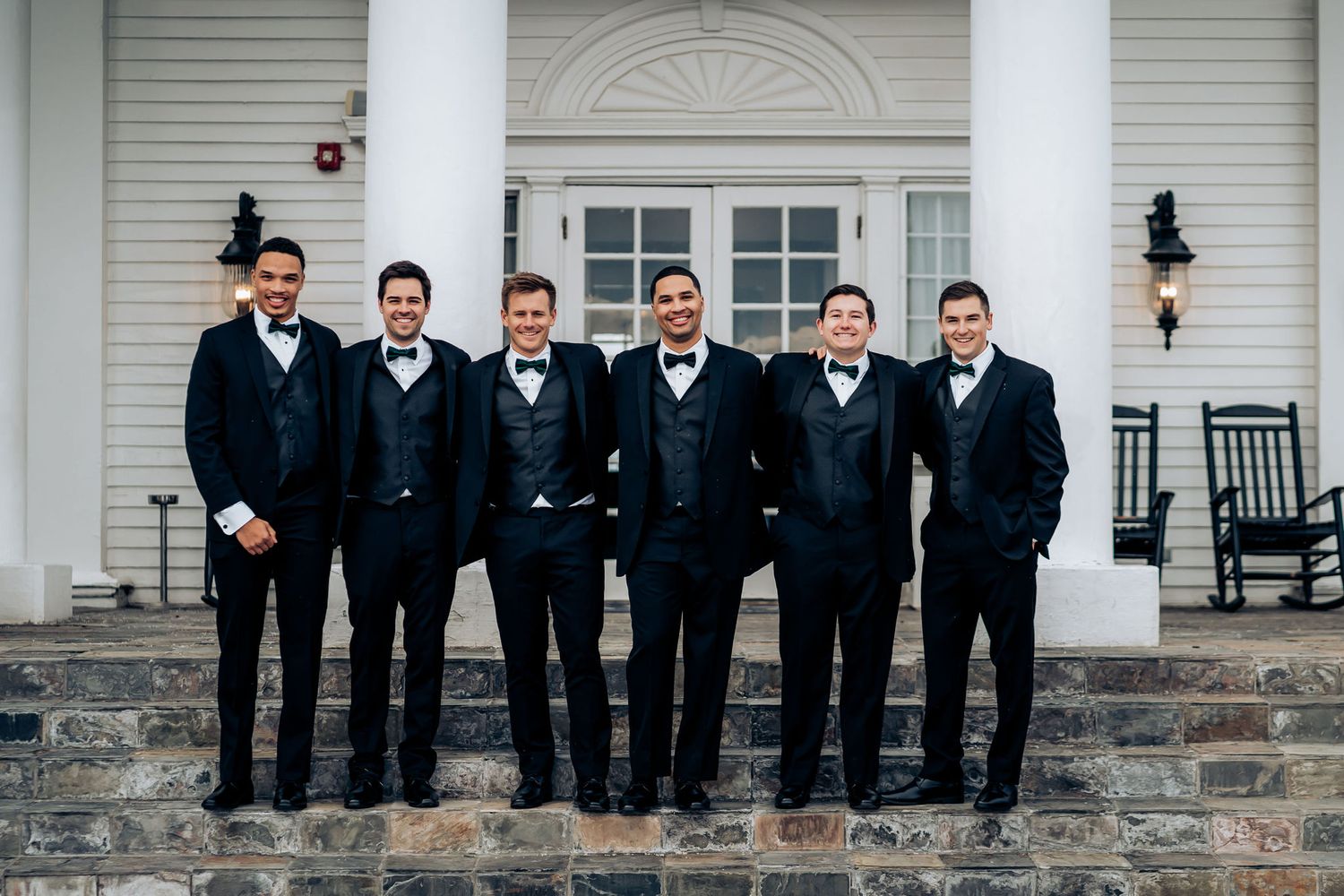 Groomsmen in black tuxedos pose on stone steps of white building.