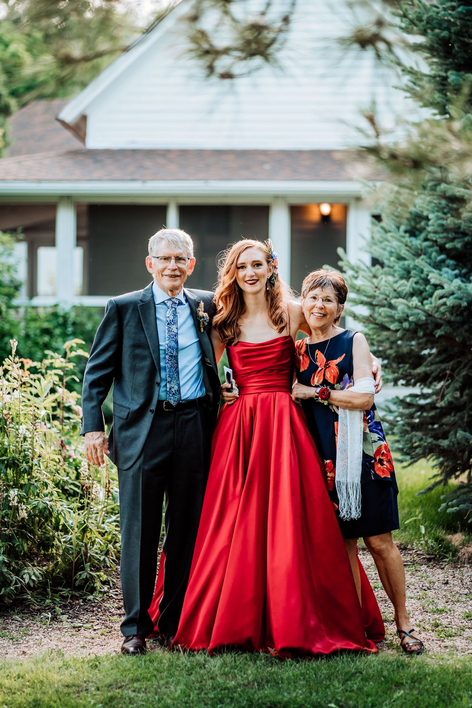 A family poses for a formal photo outside a white house with someone wearing a stunning red ball gown.