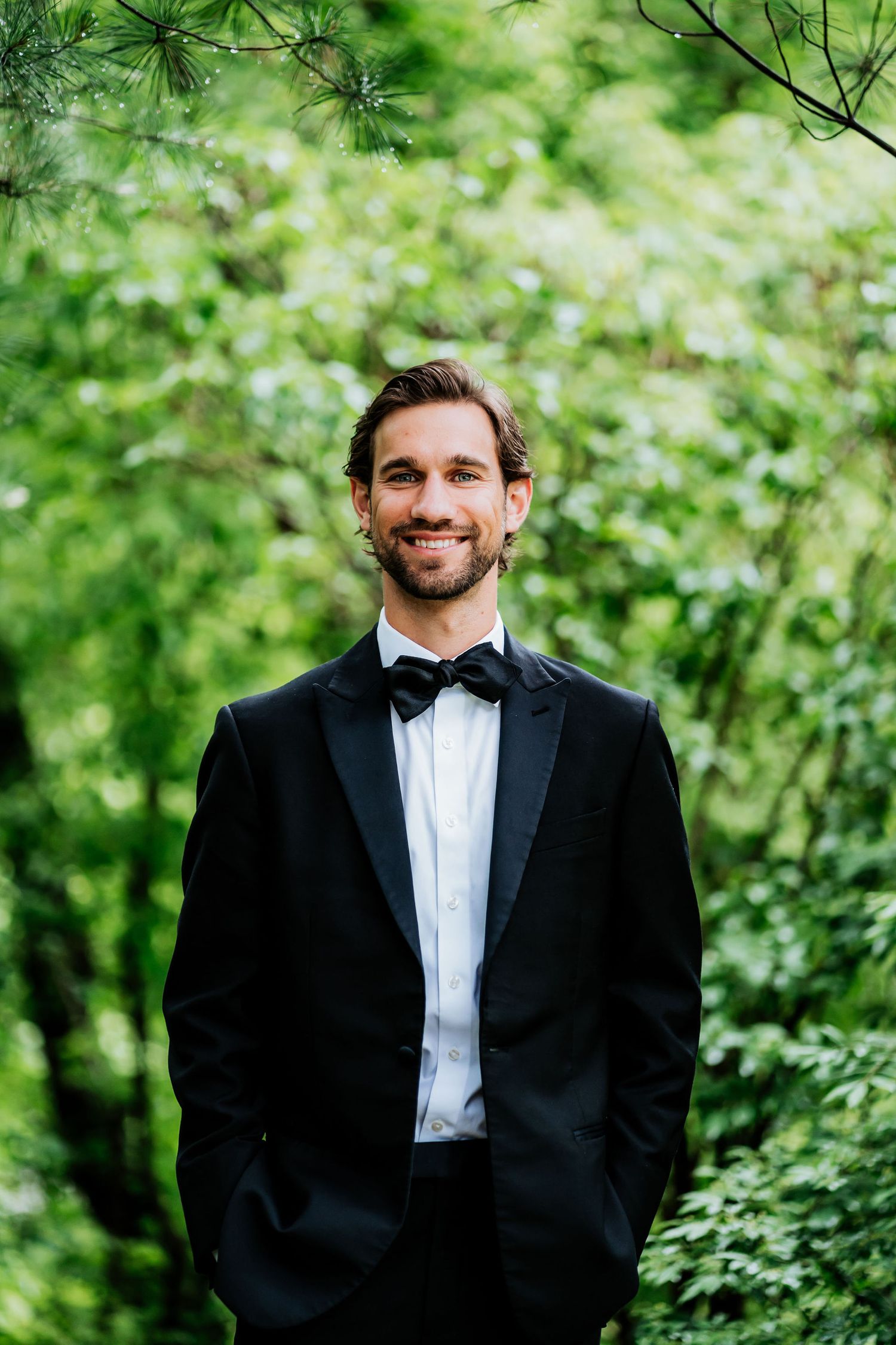 Formal portrait of someone wearing a classic black tuxedo against a green nature backdrop.