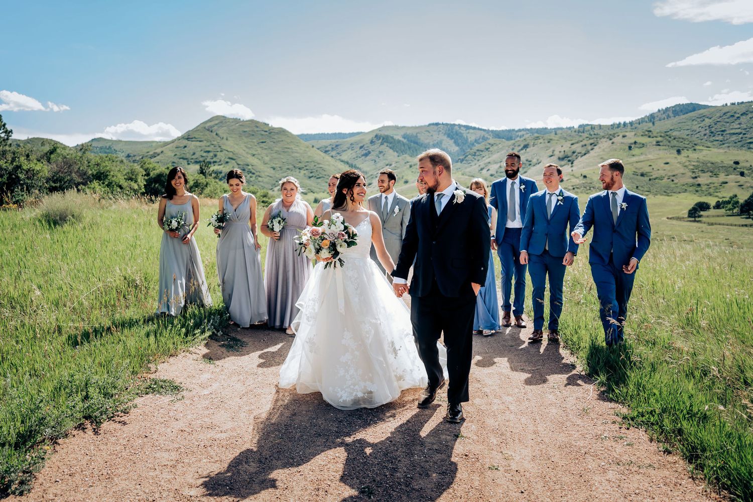 Wedding party walking along mountain trail with scenic hills in background under bright blue sky.