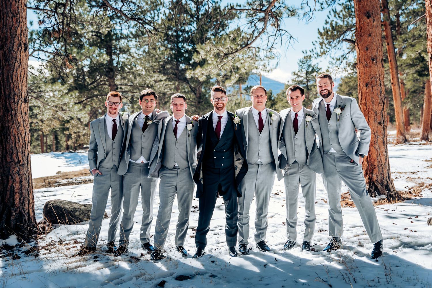 Wedding party of groomsmen in gray suits with navy-suited center figure stand in snowy pine forest.