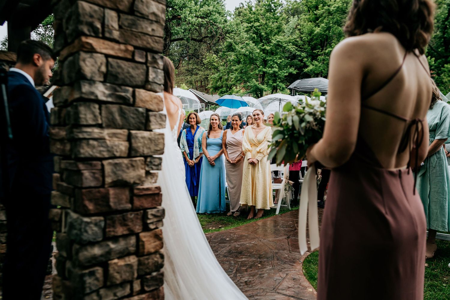 Wedding guests gather under umbrellas near a stone archway during an outdoor ceremony.
