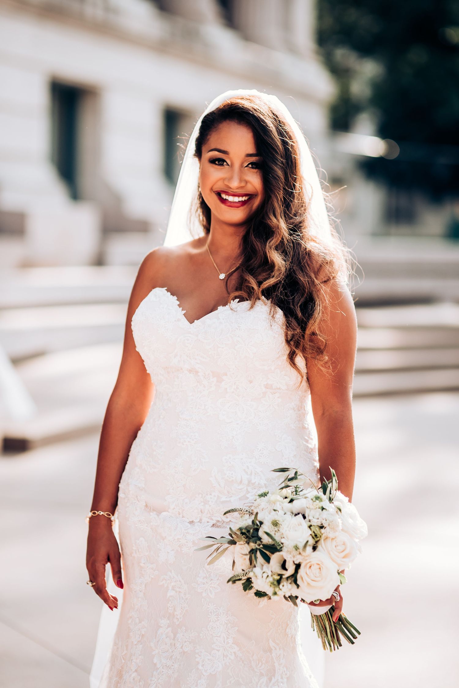 Elegant bride wearing strapless lace gown holds white floral bouquet outdoors.