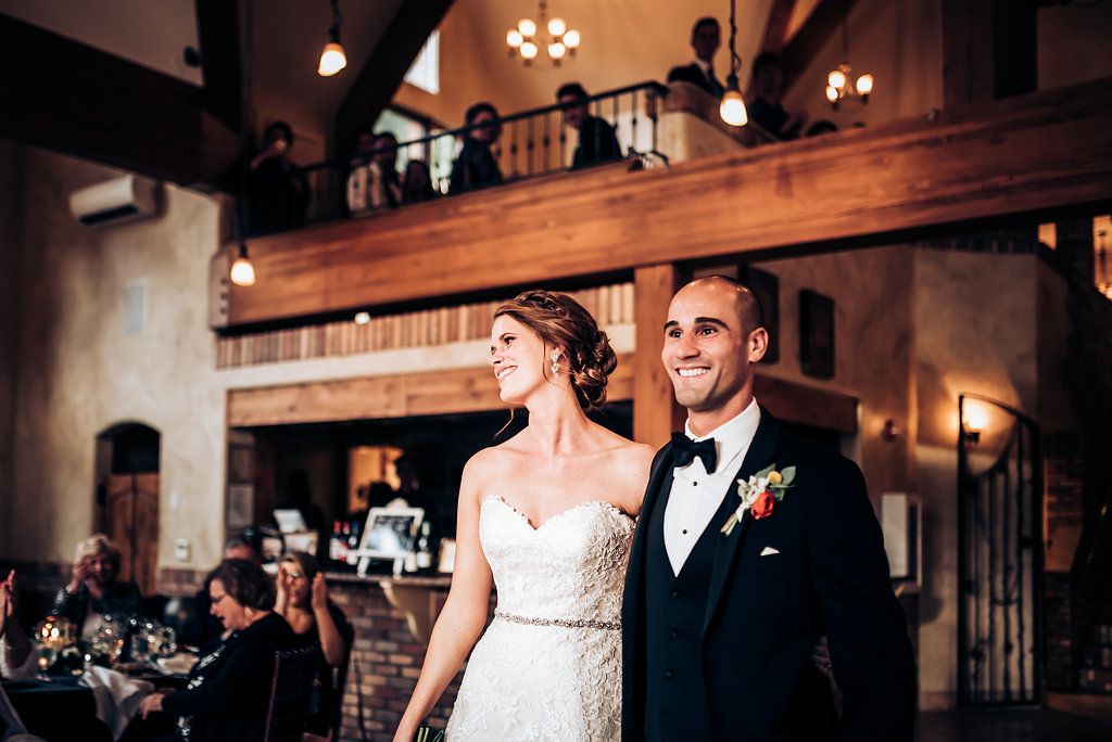 Elegant couple stands together in grand reception hall with wooden beams and warm lighting.