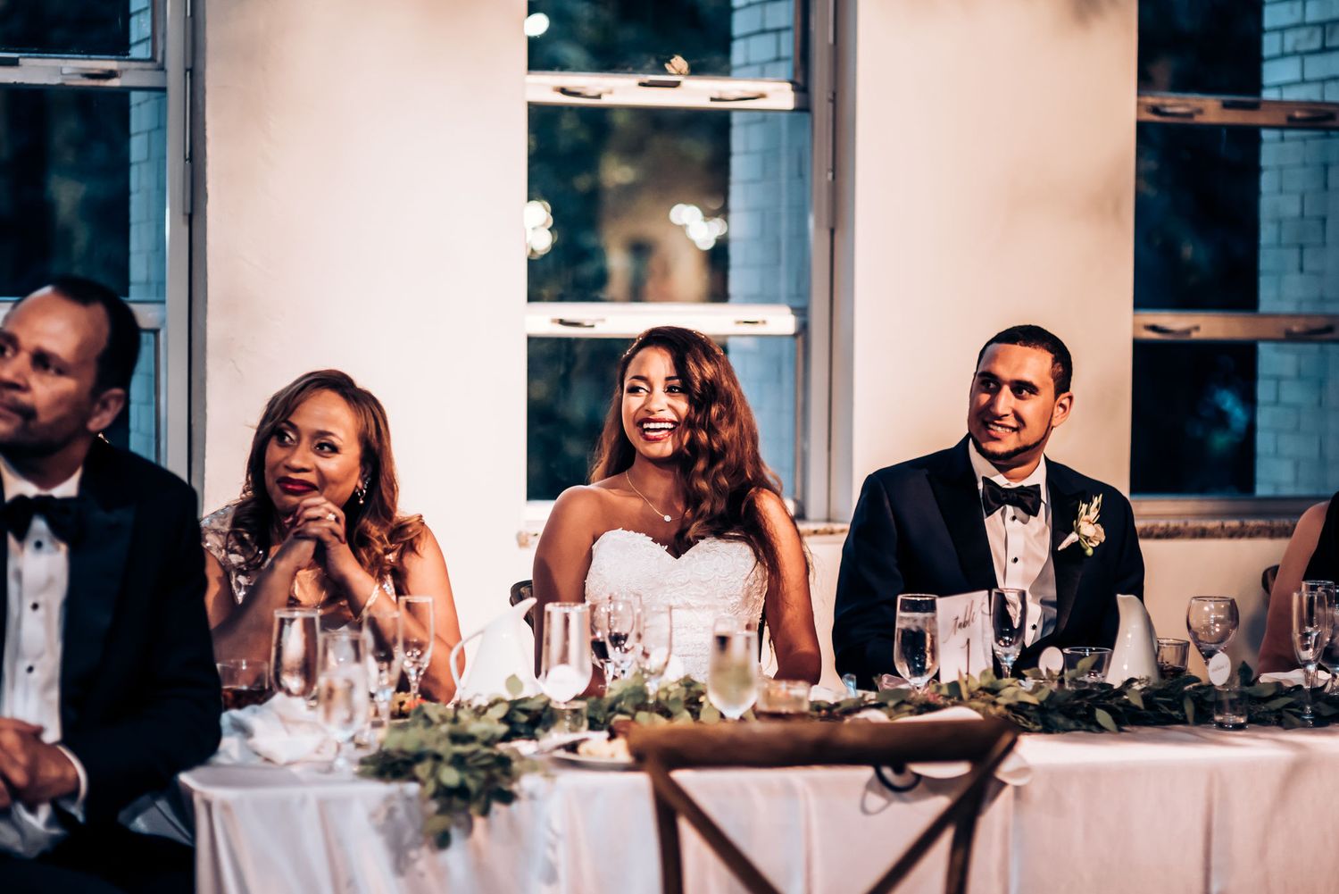 Reception head table decorated with greenery and glassware as guests share joyful moments.