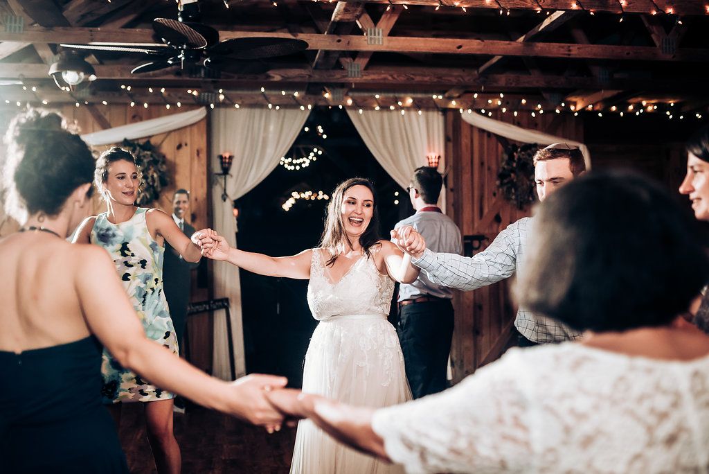 Wedding guests dance and celebrate in a rustic venue decorated with string lights and white draping.