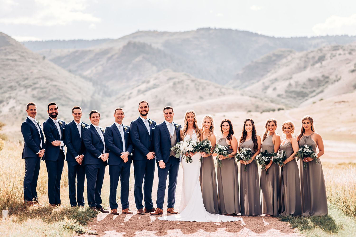 Wedding party in navy suits and gray dresses pose together against scenic mountain backdrop.