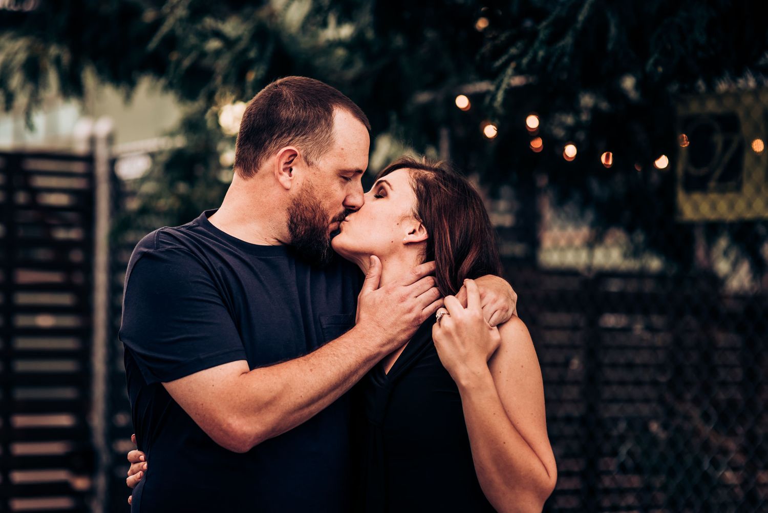 Couple sharing an intimate embrace outdoors with string lights in background.