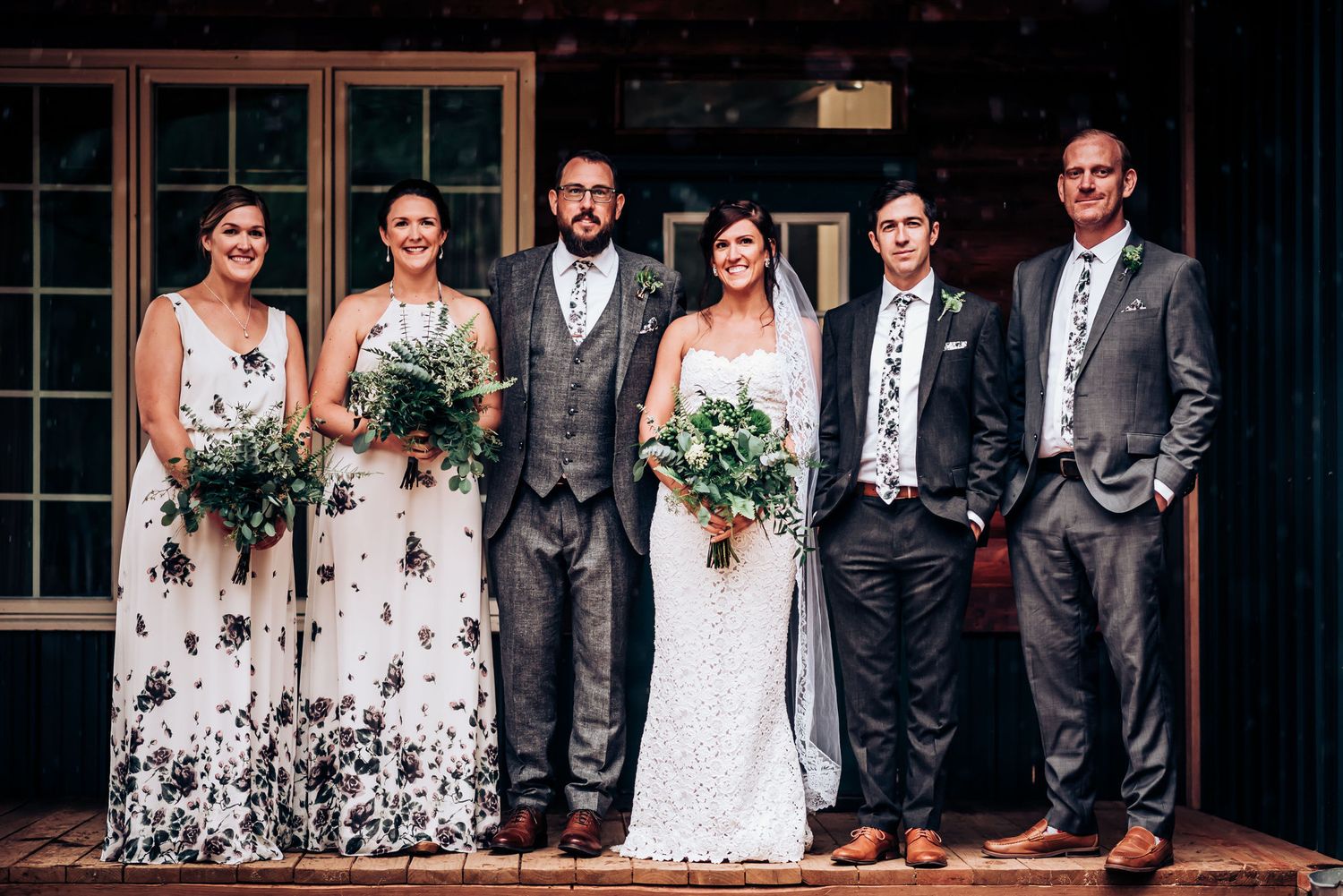 Wedding party wearing monochrome floral dresses and grey suits pose on wooden porch.