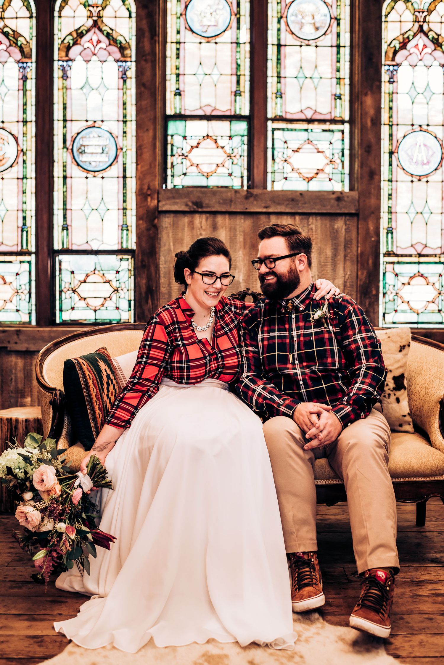 Wedding couple in coordinated plaid shirts sit together on antique couch with floral bouquet.