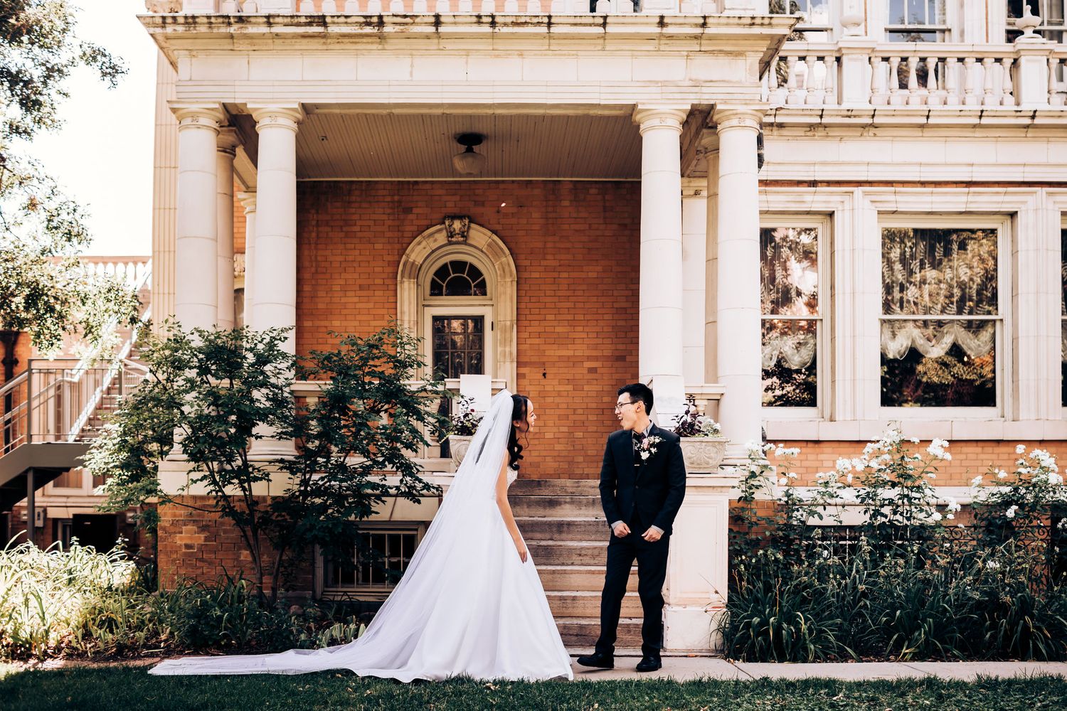 A wedding couple posing in front of a classic brick mansion with white columns.