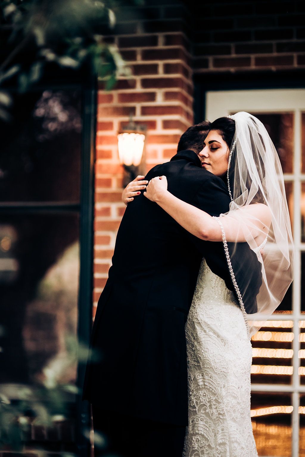 A heartfelt embrace between newlyweds beside a brick wall with a glowing lantern.