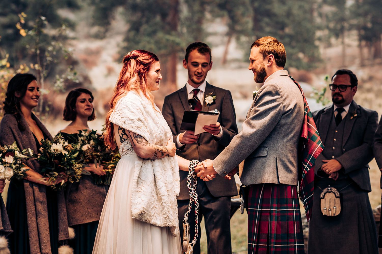 Outdoor wedding ceremony shows couple exchanging vows surrounded by wedding party in Scottish attire.