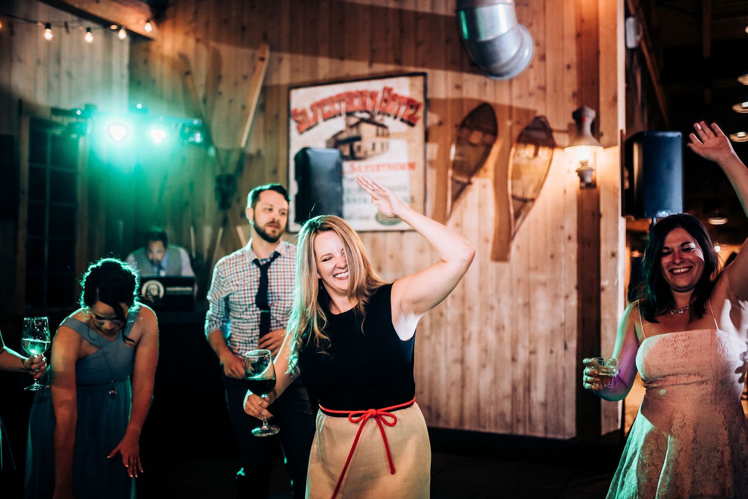 Wedding guests dance and celebrate energetically on a rustic wooden dance floor at a reception venue.
