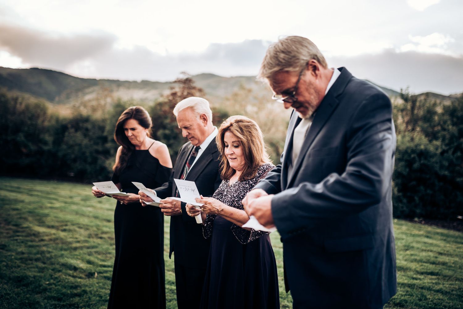 Group reading from papers during outdoor ceremony with mountain backdrop.