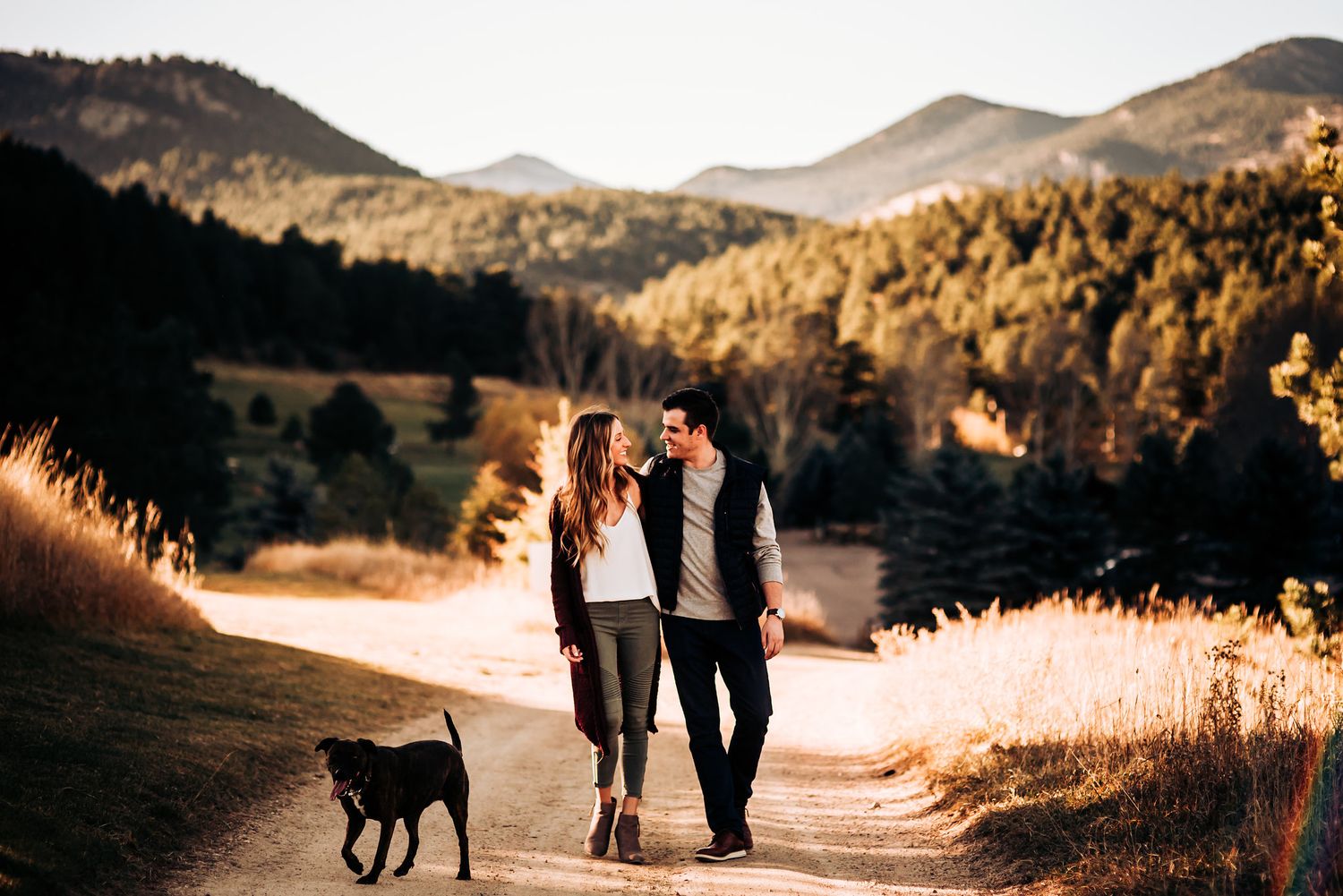 A couple walks their dog along a mountain trail with scenic peaks in the background.