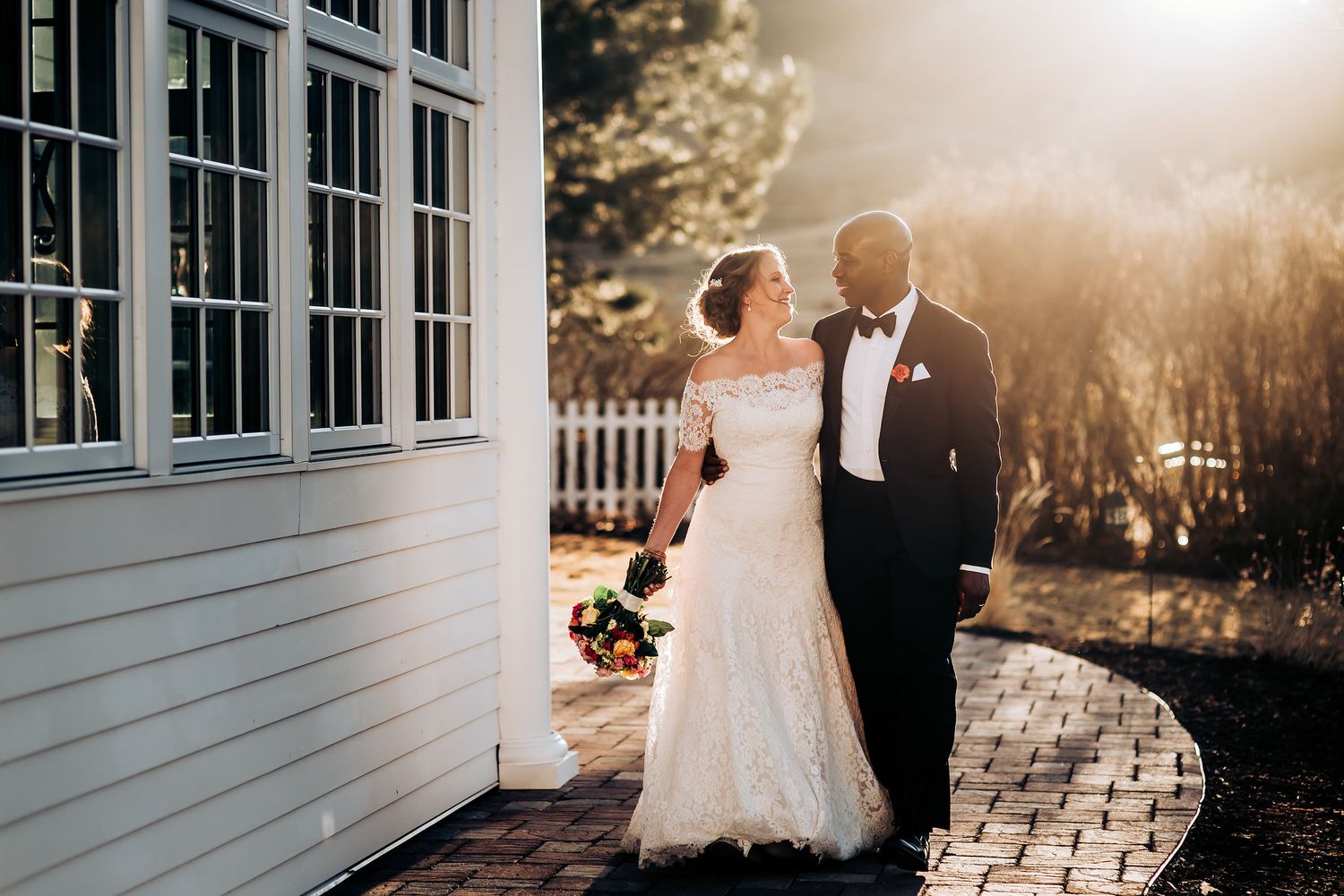 Newlyweds pose together outside white building with golden sunlight filtering through.