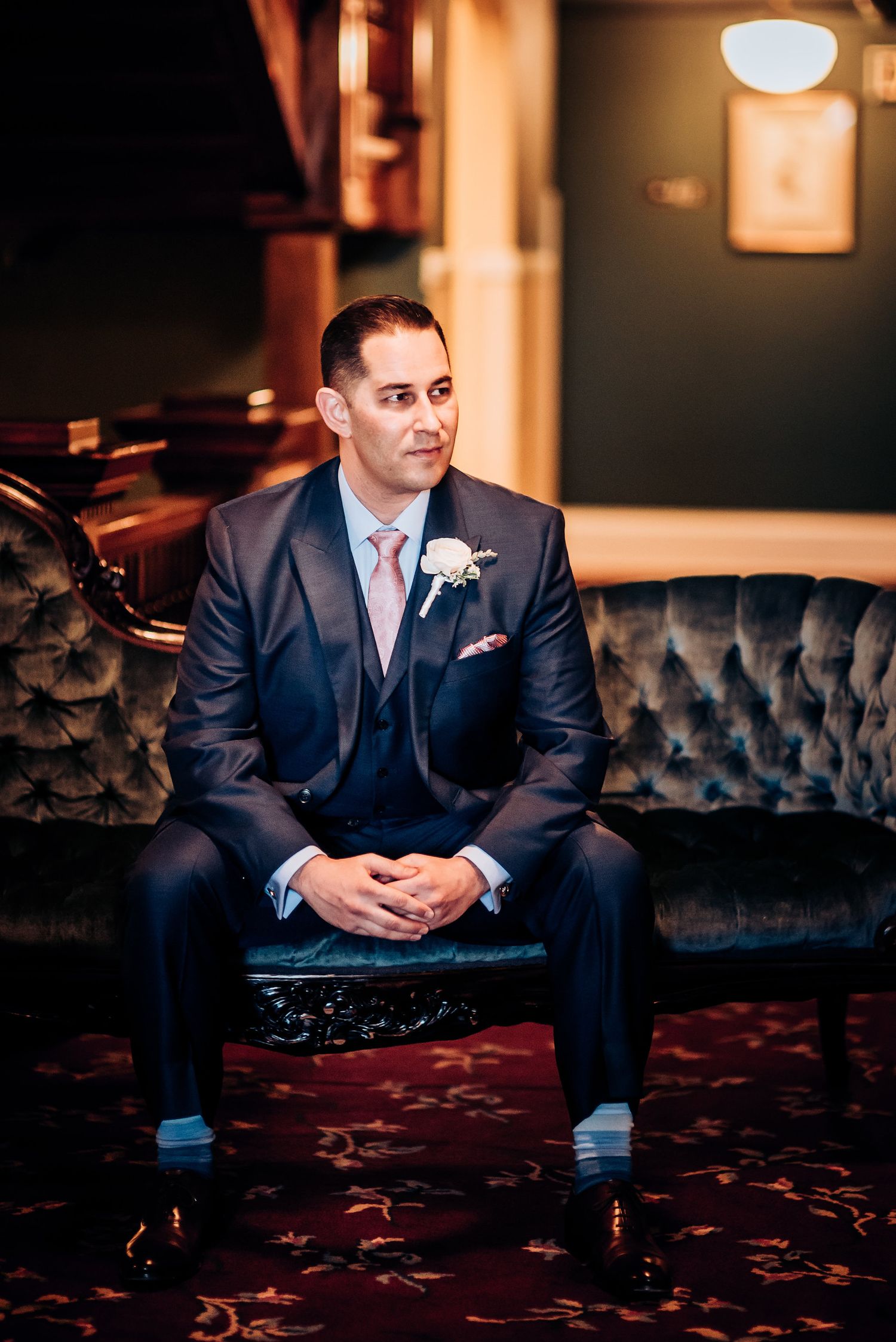 Groom in navy suit sitting on tufted velvet sofa in warm-lit room.