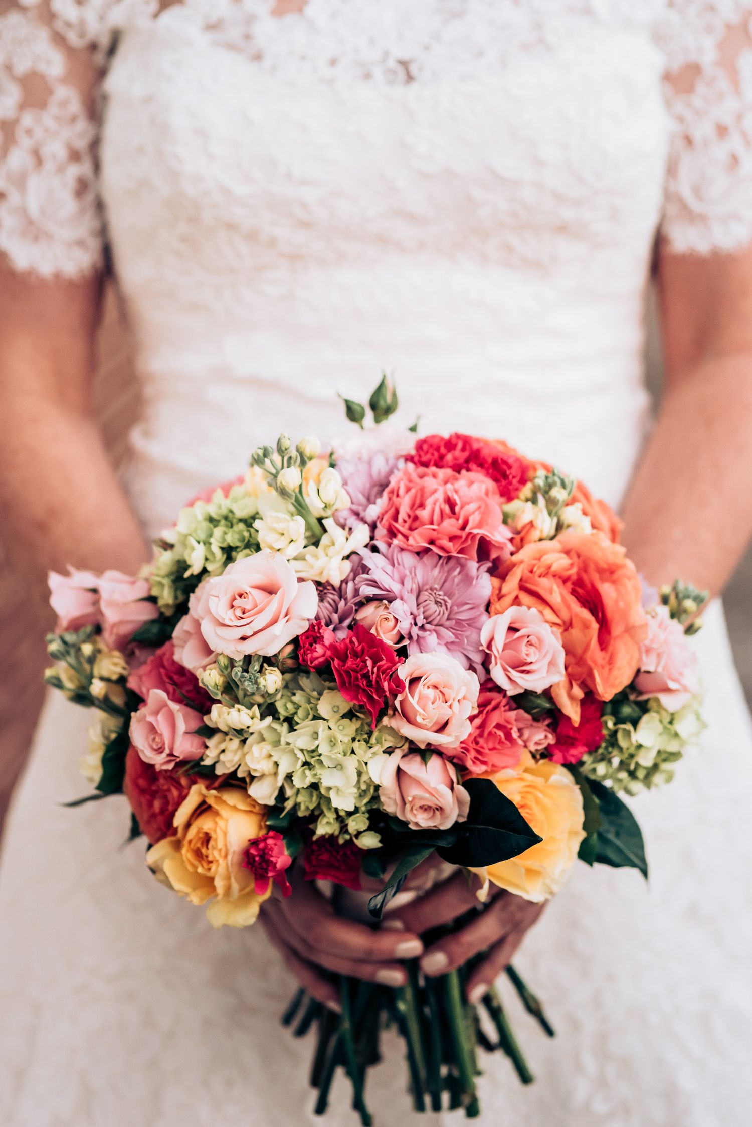 Close-up of a vibrant bridal bouquet with pink roses, peonies and mixed flowers against a white lace dress.