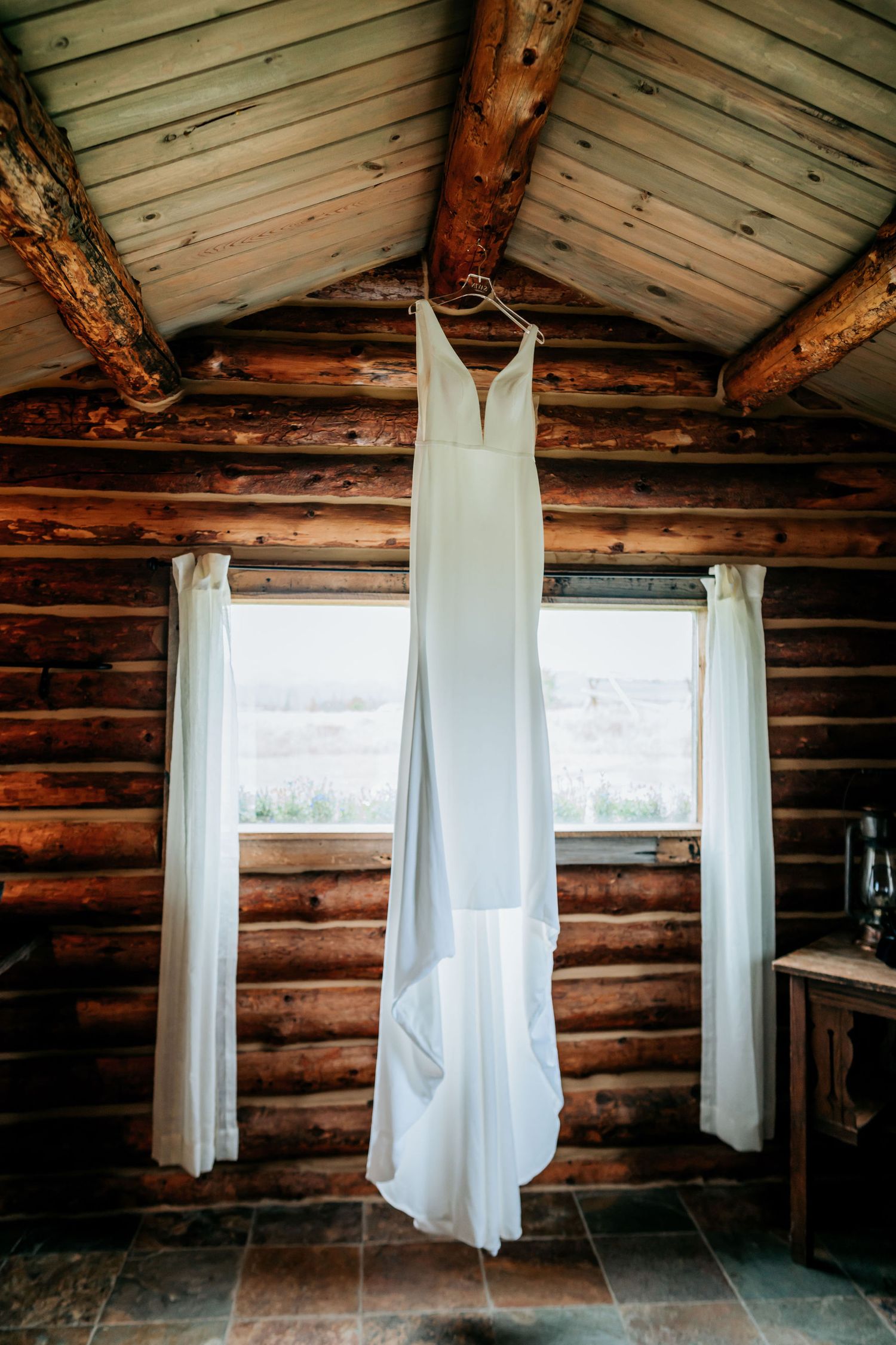 Elegant white wedding dress hanging in log cabin with natural light streaming through windows.