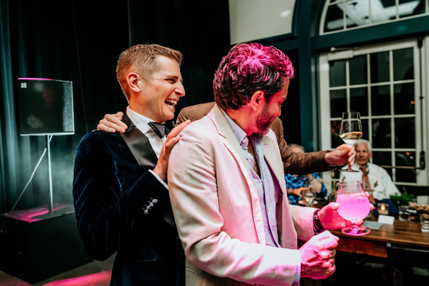 Two guests share a celebratory moment at an indoor reception with pink lighting and DJ equipment visible.