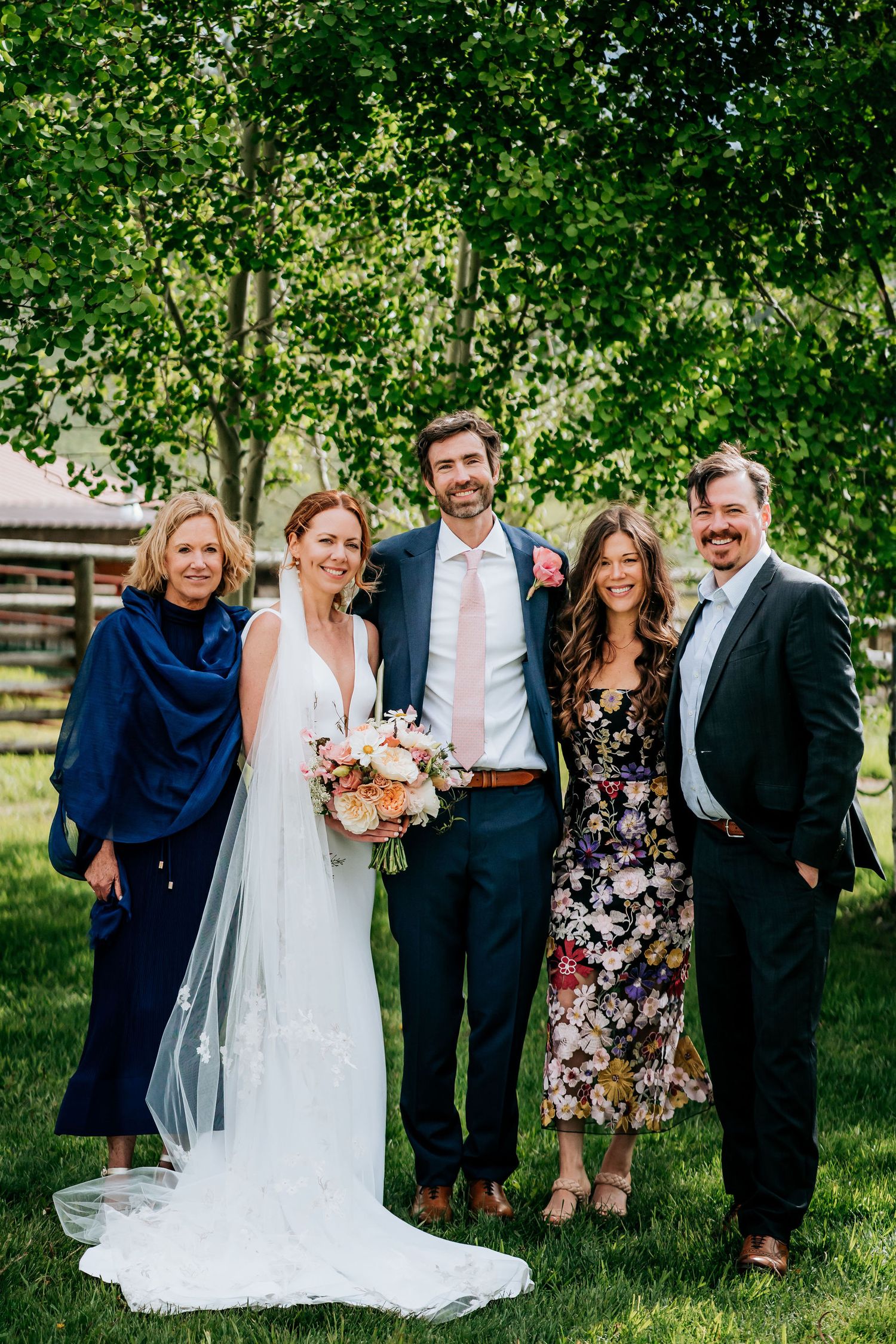 Wedding party stands together on grass with green trees in the background.