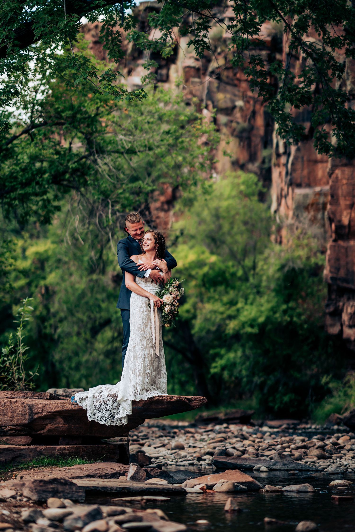 Newlyweds embrace on rocky outcrop overlooking stream with dramatic cliffs in background.